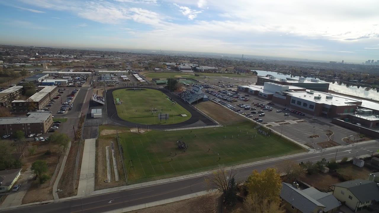 Aerial view of a sports field, track, roads, parking lots, buildings, and water beyond.