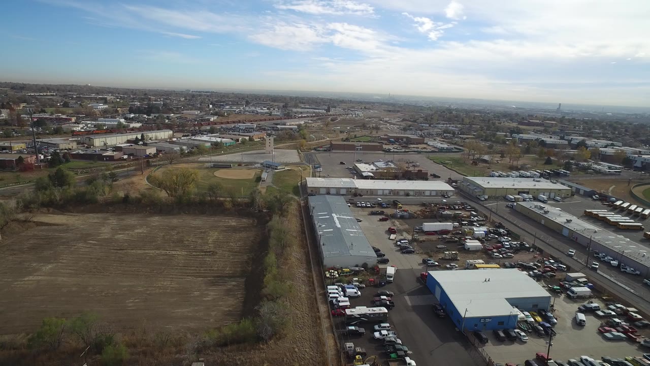 Opening aerial frame over Hidden Lake, Colorado with a brown field in front, warehouses and vehicle lots beyond it, roads crossing the midground, a distant urban horizon, and no visible people.