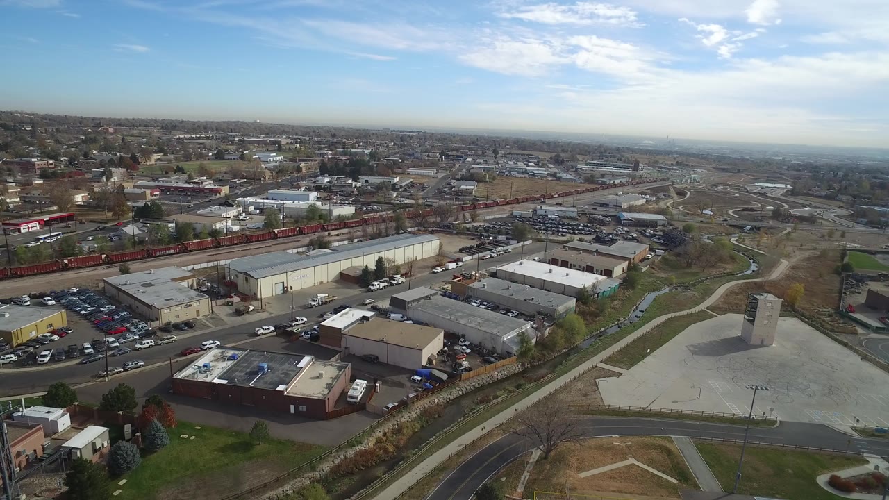 Opening aerial frame over Hidden Lake, Colorado with low-rise commercial buildings, a red freight train, open lots, and distant neighborhoods beneath a clear sky, no visible people.