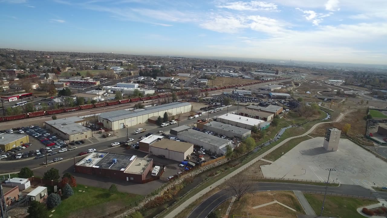 Aerial frame over Hidden Lake, Colorado with warehouse roofs in the foreground, parked vehicles along the road, a narrow drainage channel, rail cars, and a wide horizon, no visible people.