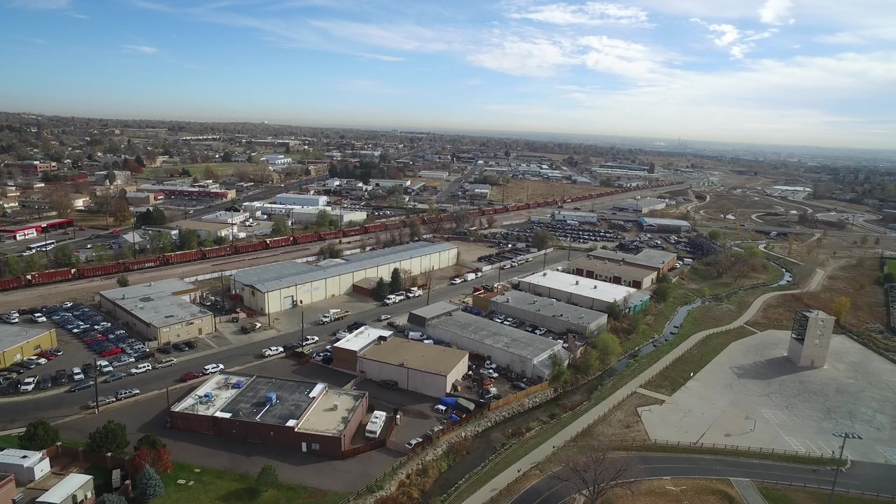 Drone-height view of commercial lots, rail line, roads, and open land near a distant cityscape.