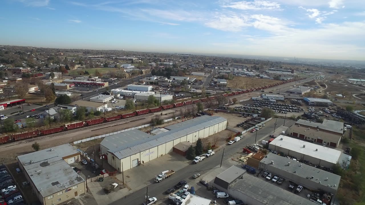 Aerial view in Hidden Lake, Colorado, with red freight cars stretching across the scene, industrial rooftops in the lower frame, distant neighborhoods, and no visible people.