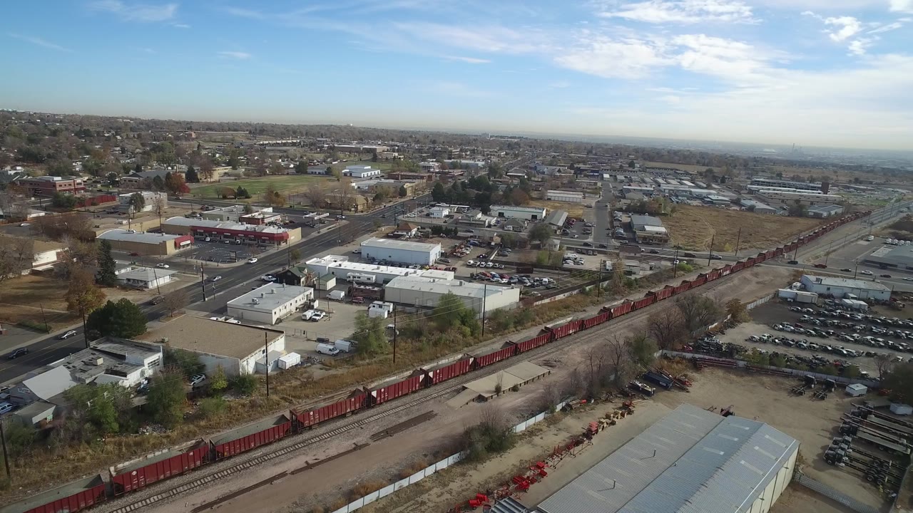 Aerial view in Hidden Lake, Colorado, with red rail cars along a diagonal track, a main road, warehouse roofs, parked cars, and no visible people.