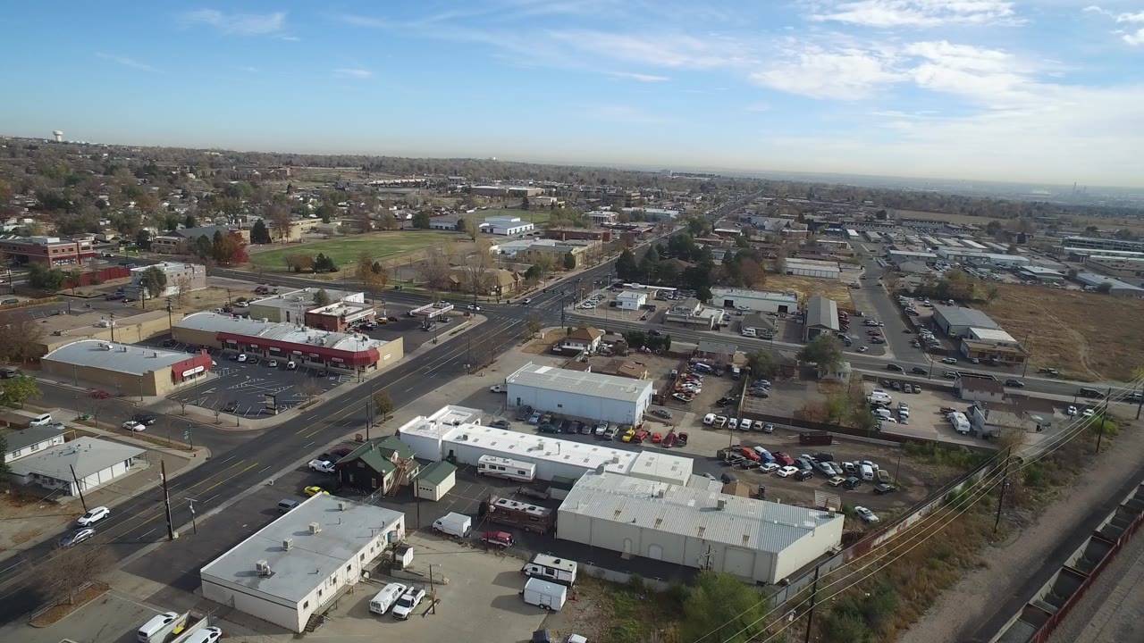 Opening aerial frame over Hidden Lake, Colorado commercial blocks, with warehouse roofs and parking lots in front, roads through the middle, distant neighborhoods on the horizon, and no visible people.