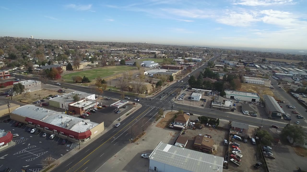 Aerial view over a suburban road intersection with commercial buildings and parked cars.