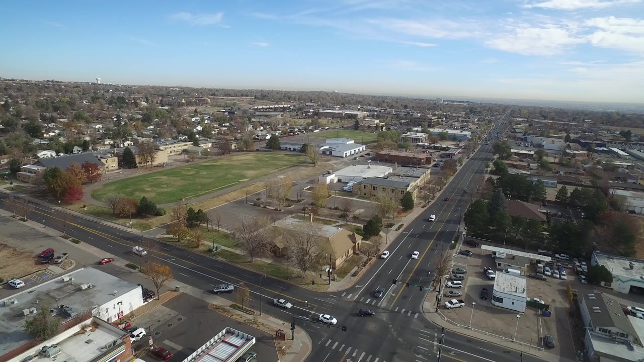 Aerial view in Hidden Lake, Colorado with the road corridor extending toward the horizon, a rectangular green field on the left, small commercial or civic buildings, parking areas, and scattered cars, with no clearly identifiable people visible.
