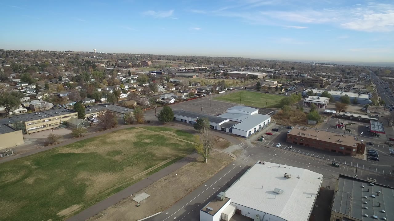 Opening aerial frame over Hidden Lake, Colorado, with a grassy field in front, low commercial buildings and parking lots below, distant houses and no visible people.