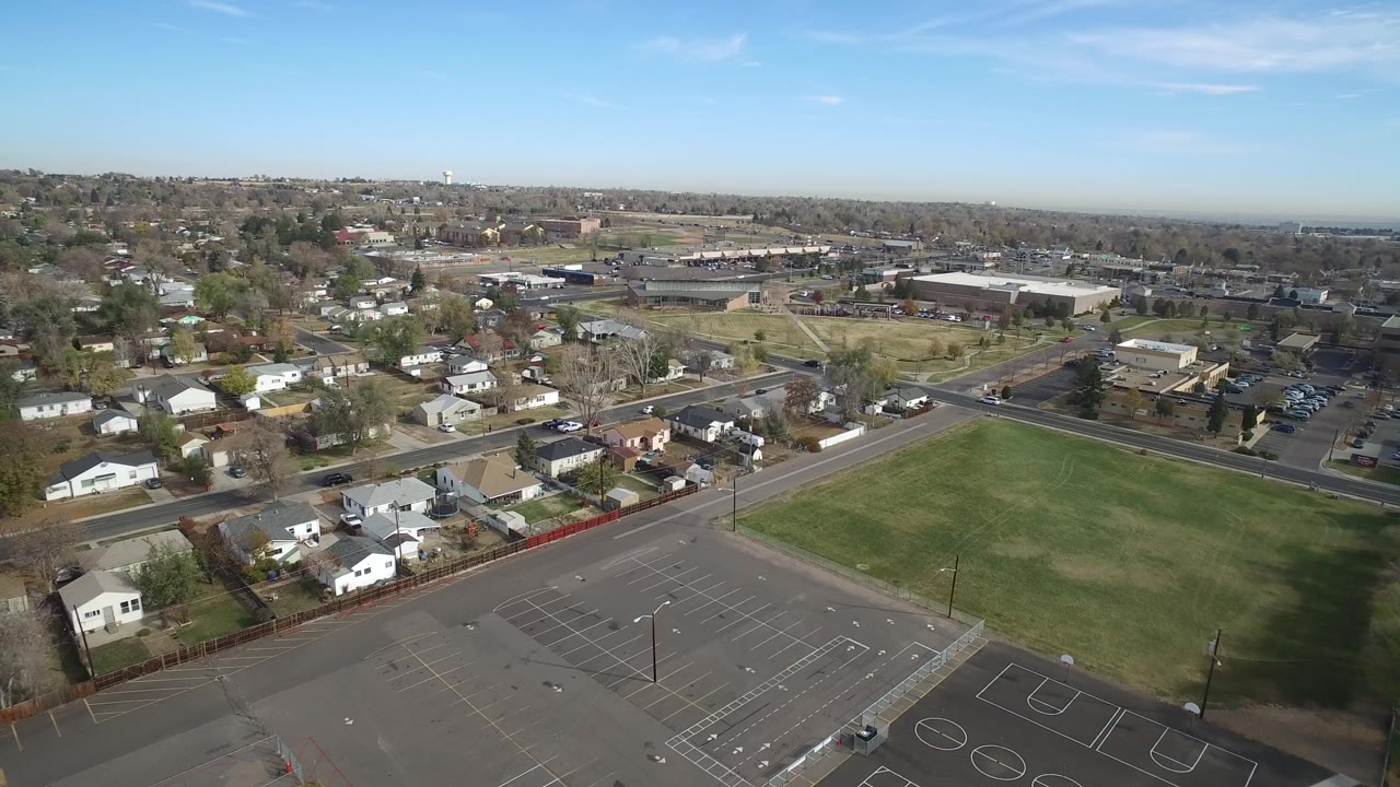 Aerial view over a green field, parking lot, roads, homes, and low buildings.