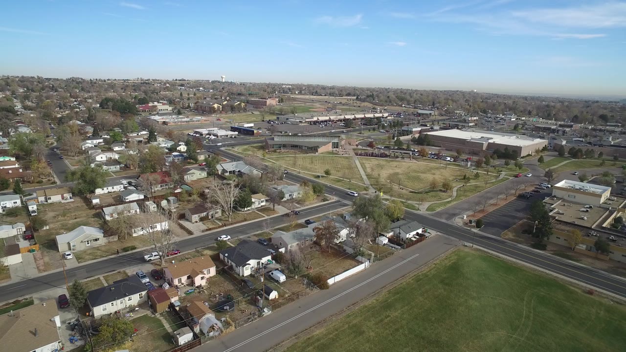Aerial view of suburban streets, houses, grass field, and low buildings under a clear sky.