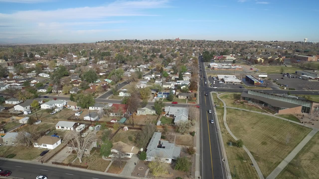Aerial frame over Hidden Lake, Colorado showing residential houses in the foreground, a straight avenue at right, retail lots beyond, and no clearly visible people.