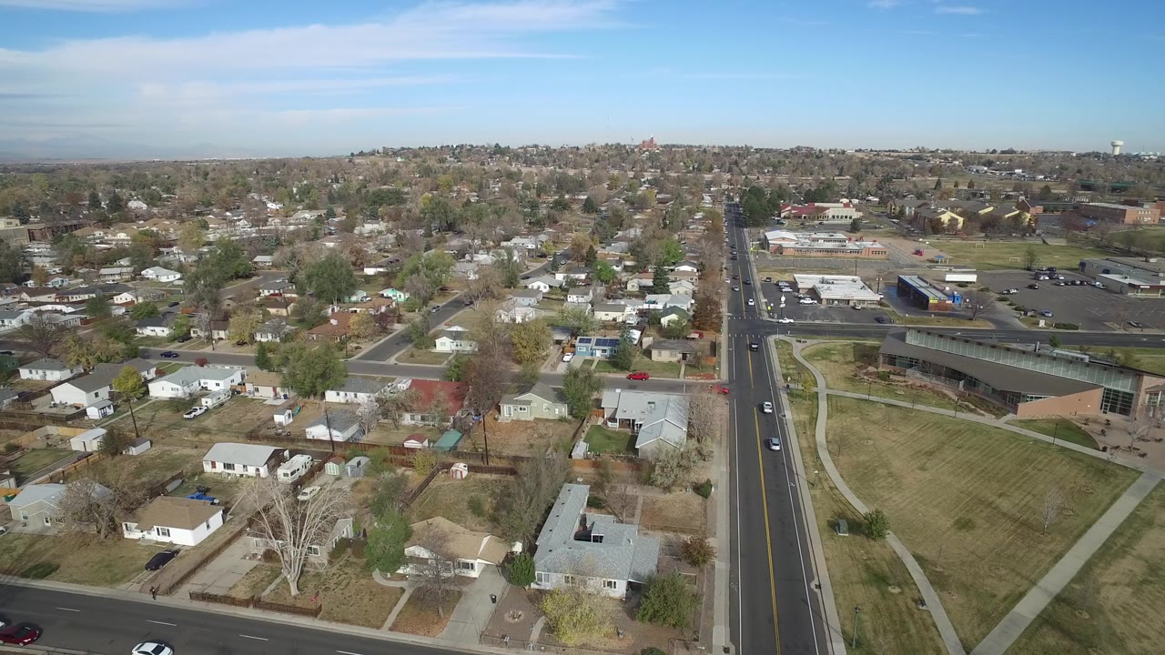 Aerial view in Hidden Lake, Colorado looking across low homes and bare trees toward a main avenue, parking areas, and low storefront buildings in bright daylight.