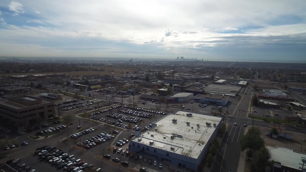 Aerial view over commercial rooftops, parking lots, roads, and a distant skyline under clouds.