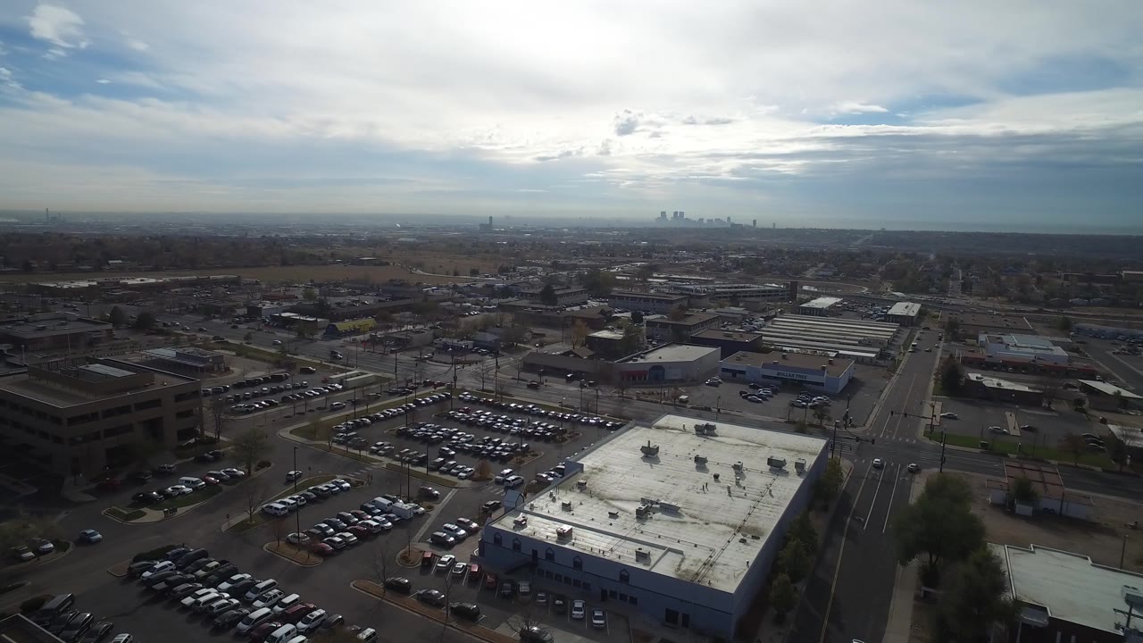 Aerial frame in Hidden Lake, Colorado showing a white flat-roof building, rows of parked cars, a straight road leading toward the horizon, distant towers, and no visible people.