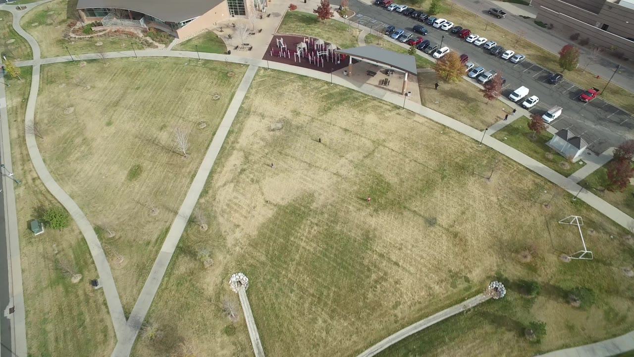 Aerial view in Hidden Lake, Colorado, with pale grass fields crossed by sidewalks, a tan building near the top, parked cars to the right, and tiny figures too small to identify.