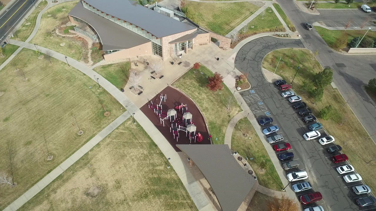 Overhead frame in Hidden Lake, Colorado showing a tan building beside a red play or exercise surface, a grassy field, parked cars, and surrounding roads with no visible people.