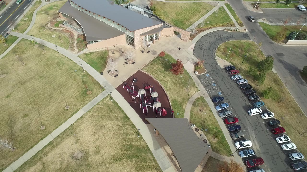 High drone view over a grassy field, building, red play area, parking row, and road intersection.