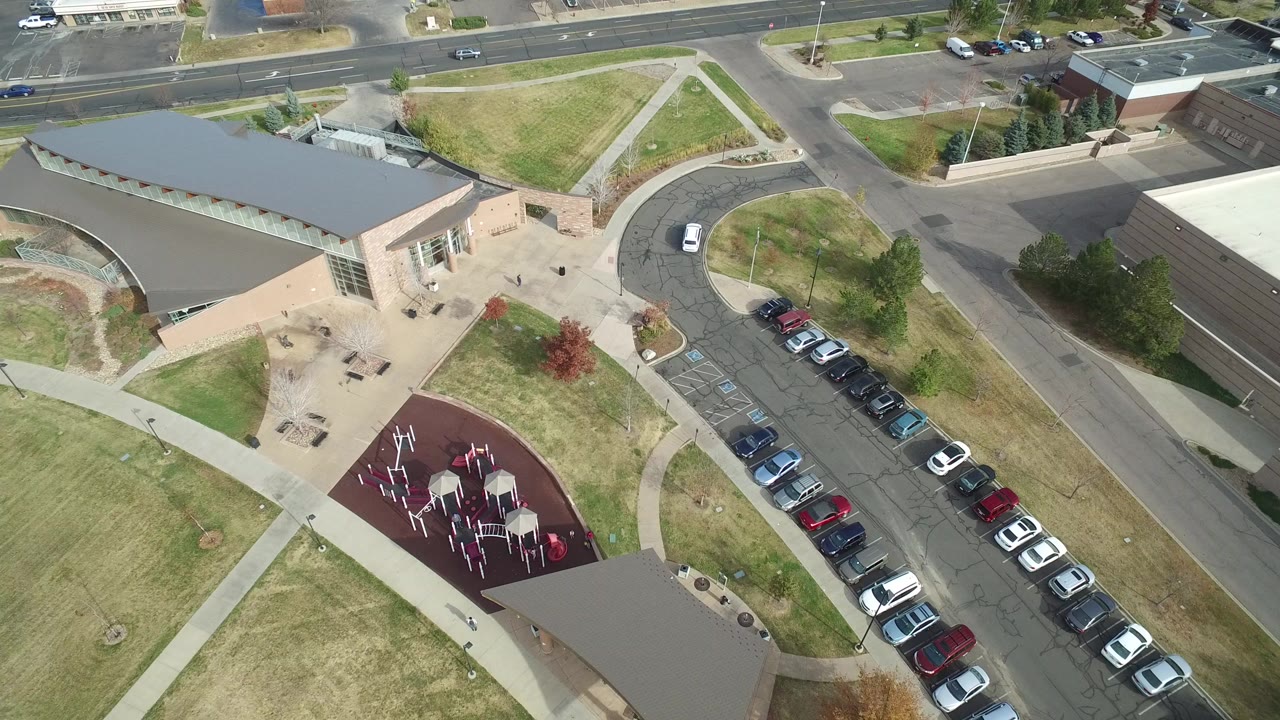 High aerial frame over Hidden Lake, Colorado showing a tan building, red playground area, grassy lawns, curving drive, filled parking row, and no visible people.