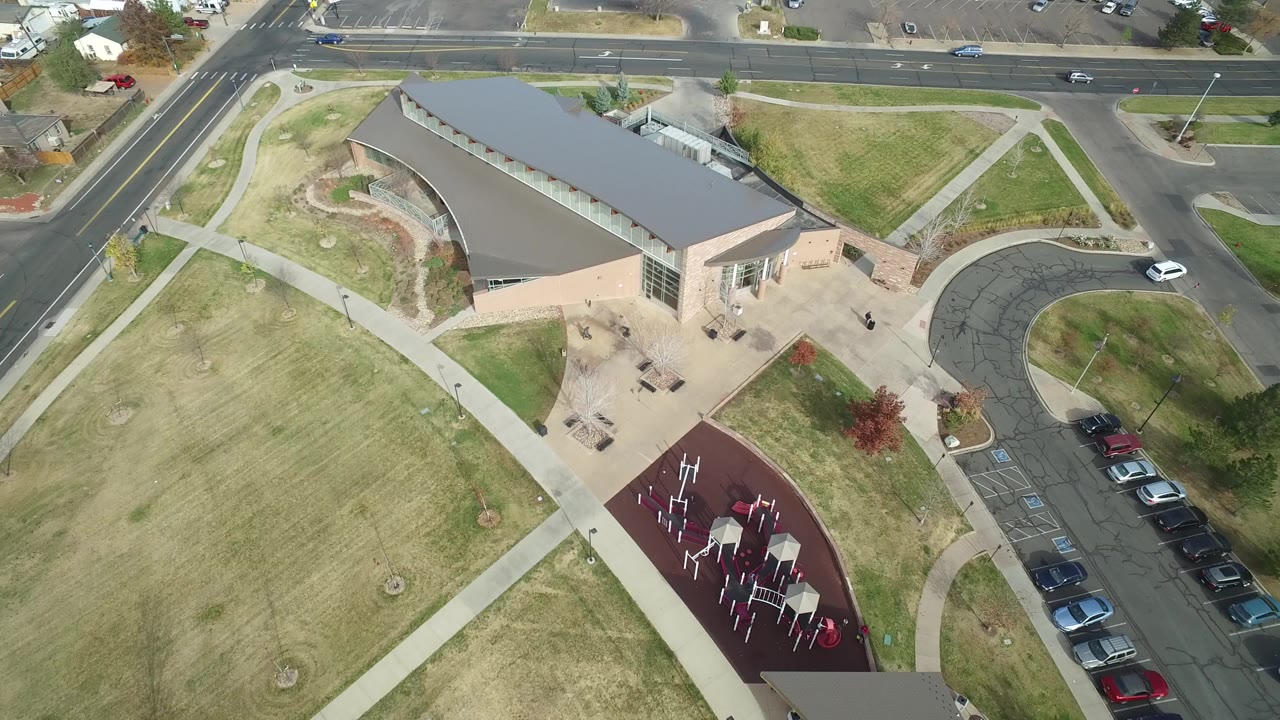 Oblique aerial view of a modern building, lawns, sidewalks, parked cars, and a red playground surface.