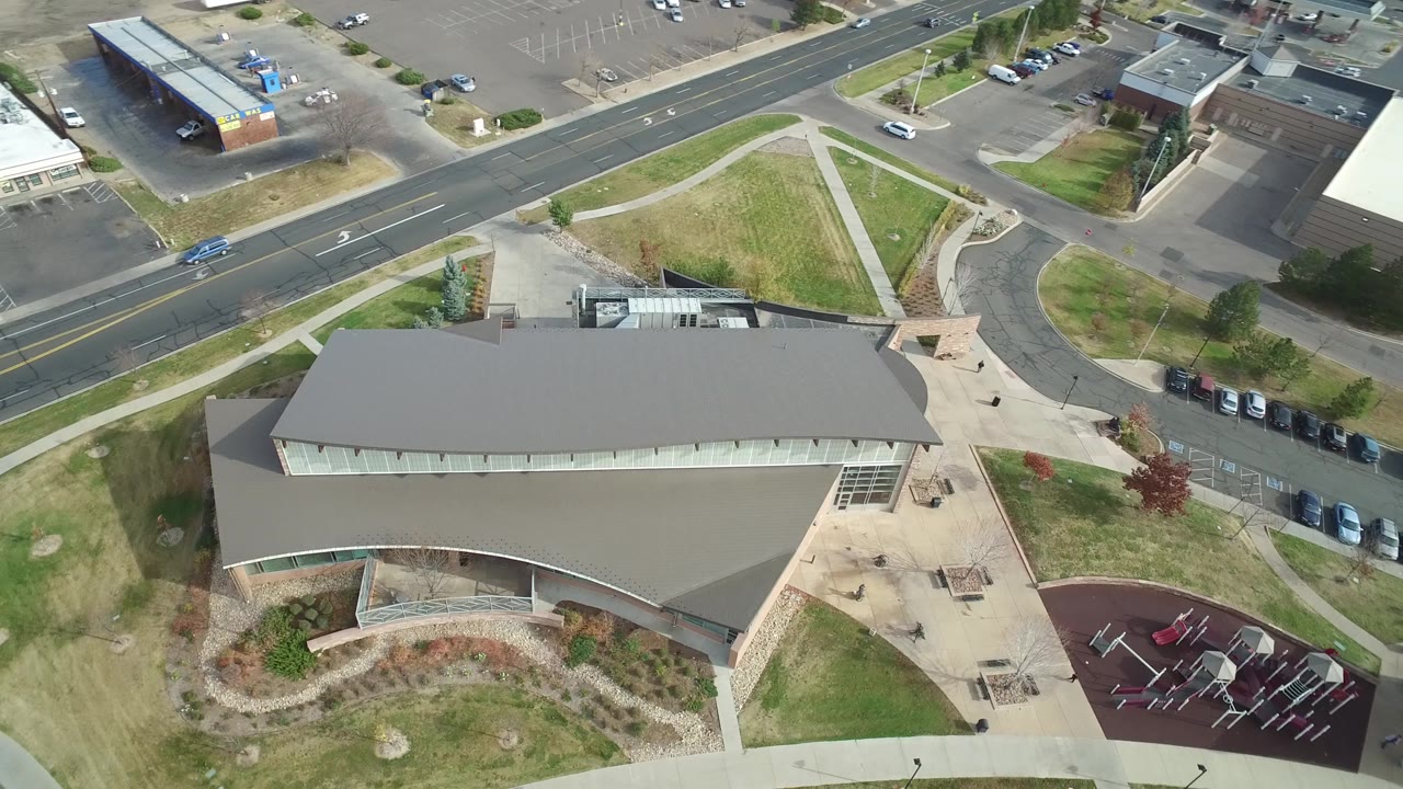 Aerial frame over Hidden Lake, Colorado showing the campus roof centered low in frame, the playground at right, diagonal sidewalks, parked cars, roads, and no visible people.