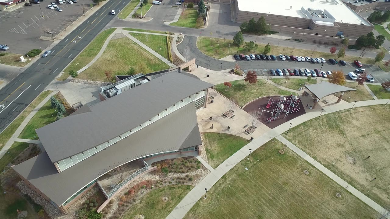 Overhead drone view of a large building, sidewalks, lawn, road, parking, and playground.