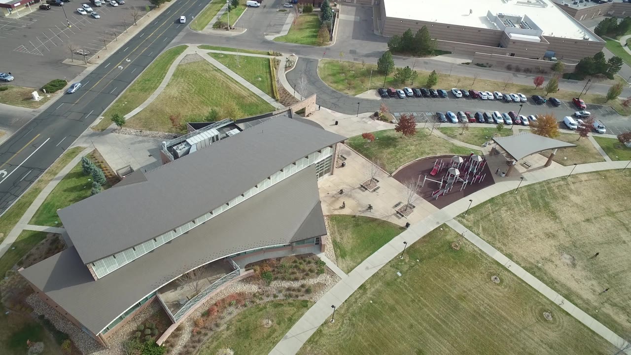 Hidden Lake, Colorado overhead frame with the roof filling more of the lower view, sidewalks crossing lawns, rows of parked cars, a road, and the playground visible beyond, with no visible people.