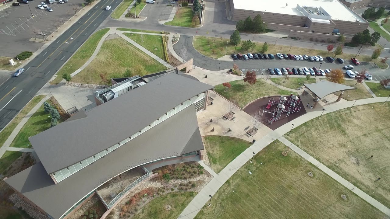 Aerial frame in Hidden Lake, Colorado with the building roof at the lower left, road lanes along the left edge, parking and another building at the top, and a playground to the right, with no visible people.