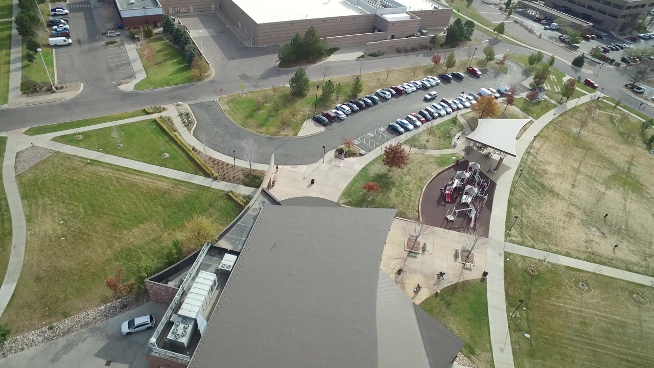 Opening aerial frame over Hidden Lake, Colorado, showing office roofs at the bottom, grass lawns crossed by sidewalks, parked cars near the top, nearby roads, and no clearly visible people.