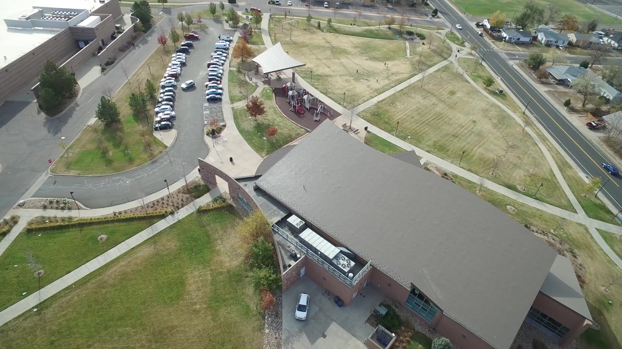 Aerial view in Hidden Lake, Colorado with the large roof filling the lower right, a full parking row at left, a small covered play area near the center, and houses beyond the road.