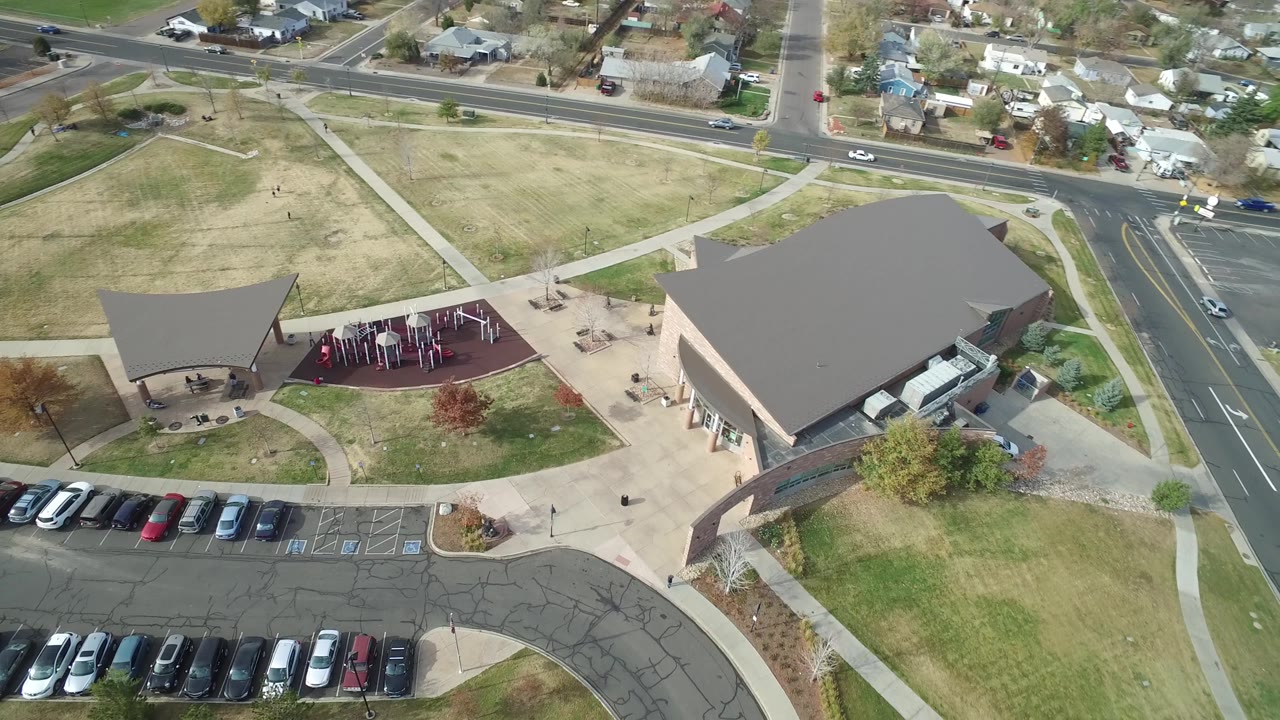 Hidden Lake aerial frame with the large roof near center, the playground and pavilion to the left, parked cars along the bottom edge, and neighborhood homes beyond the road with no visible people.
