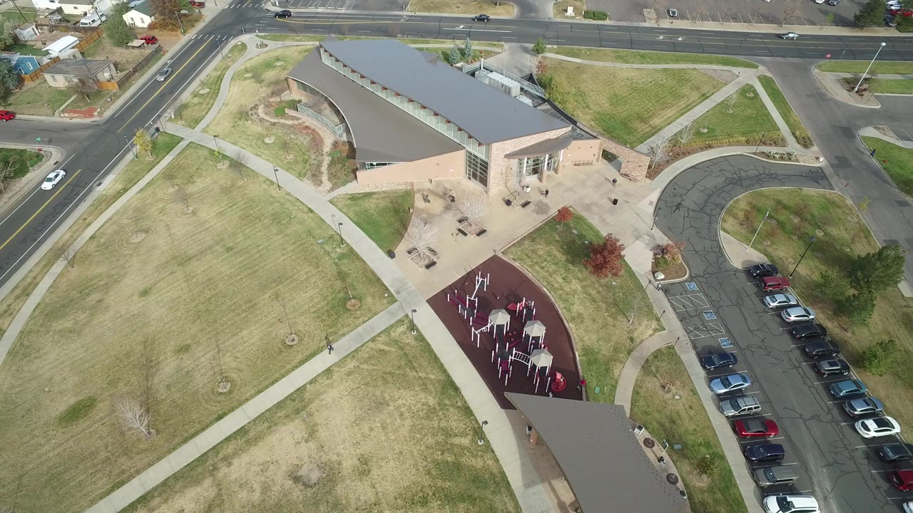 At 0:00, an overhead frame in Hidden Lake, Colorado shows a tan building with a gray roof beside dry lawns, crossing sidewalks, a red playground area, a curved road, and parked cars, with no visible people.