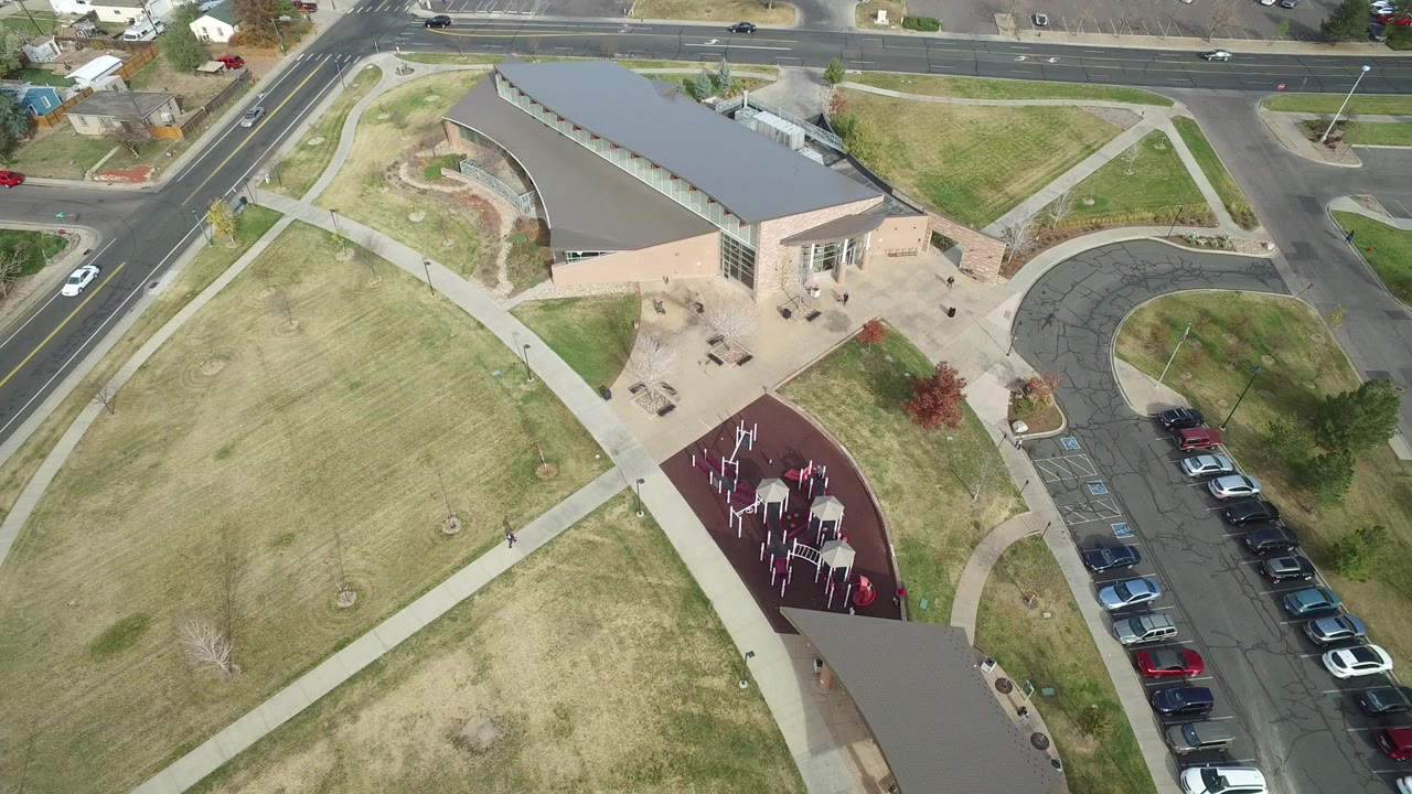 At 0:01, the aerial view holds over Hidden Lake, Colorado with the building shifted slightly left in frame, open lawn filling the lower left, the red play surface near the lower center, and parked cars along the right edge, with no visible people.
