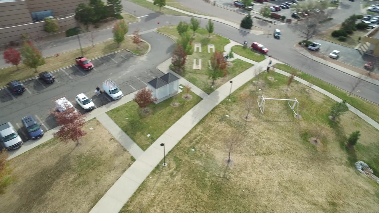 High aerial frame over Hidden Lake, Colorado showing dry grass, intersecting sidewalks, parked cars, a road curve, low buildings, and no recognizable people visible.