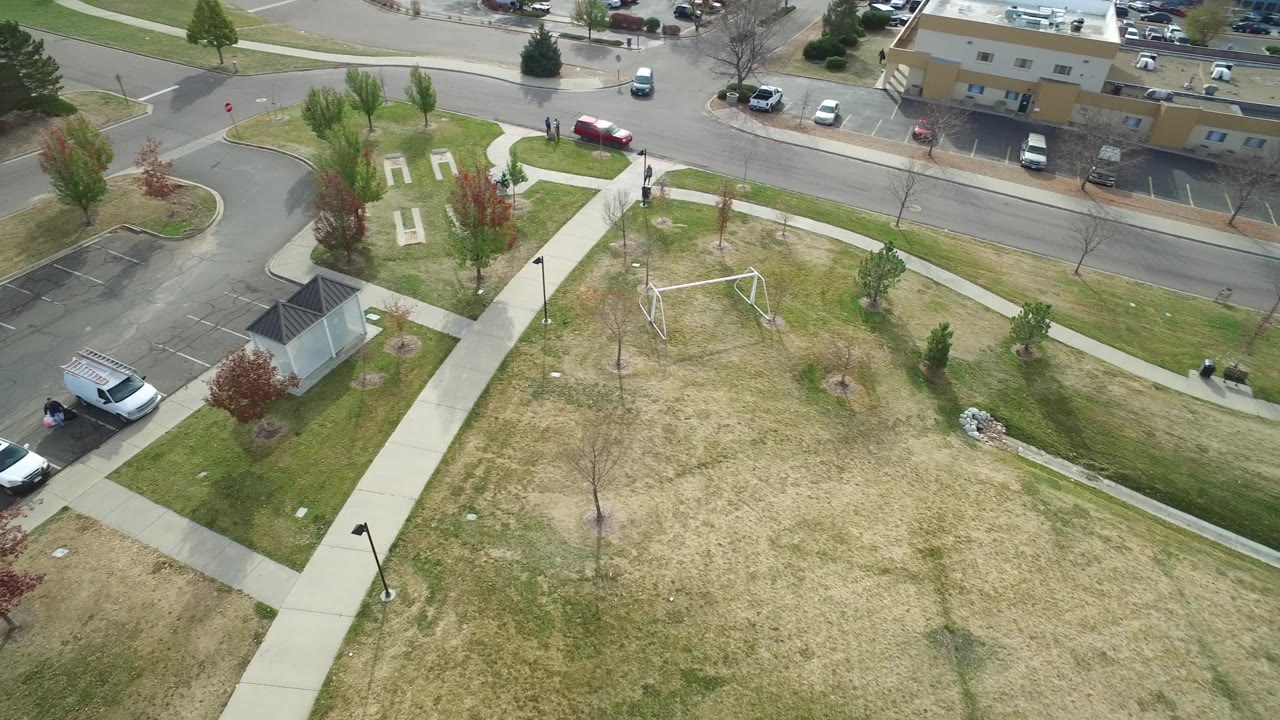Aerial view of grass, sidewalks, a white frame structure, cars, and low buildings.