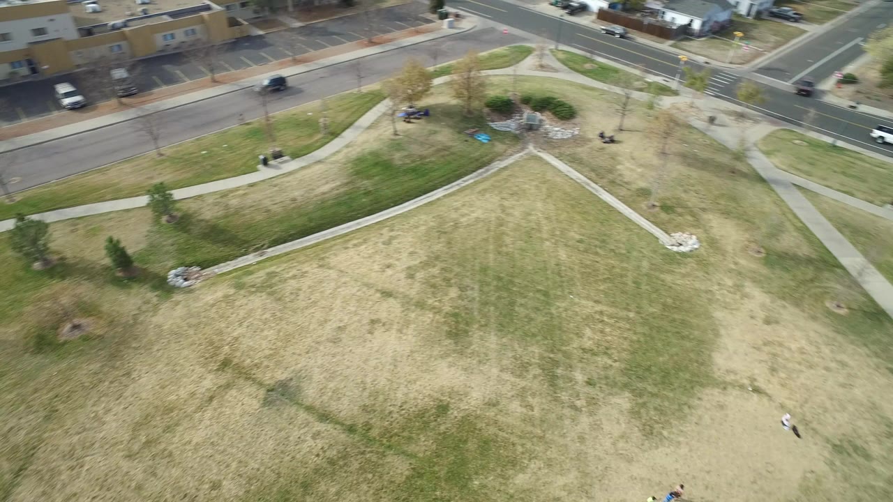 High aerial view in Hidden Lake, Colorado, showing a patchy grass lawn crossed by sidewalks with a road, parked and moving vehicles, and no clearly identifiable people visible.