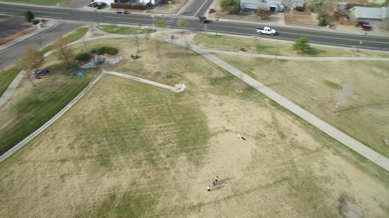 Overhead view of a grassy field, sidewalks, roadside, and distant people.