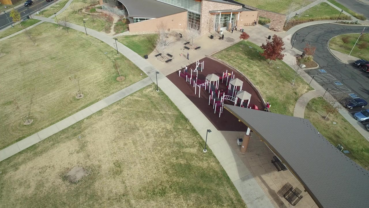 Aerial view of lawns, sidewalks, playground, building entrance, and parked cars.