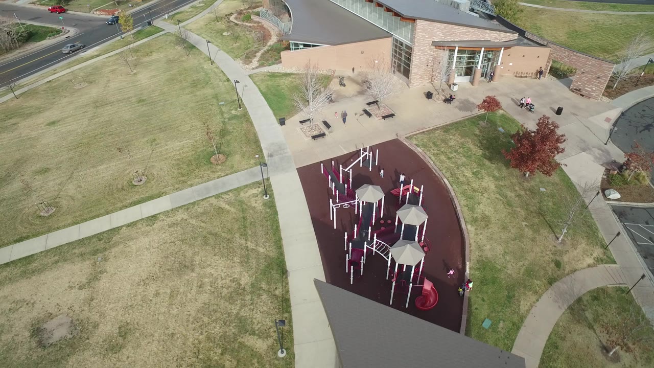 Aerial frame over Hidden Lake, Colorado shows the gray roof of a yellow building, pale lawns, a curving sidewalk, a roadside with cars, and part of a red playground area with small visible people.