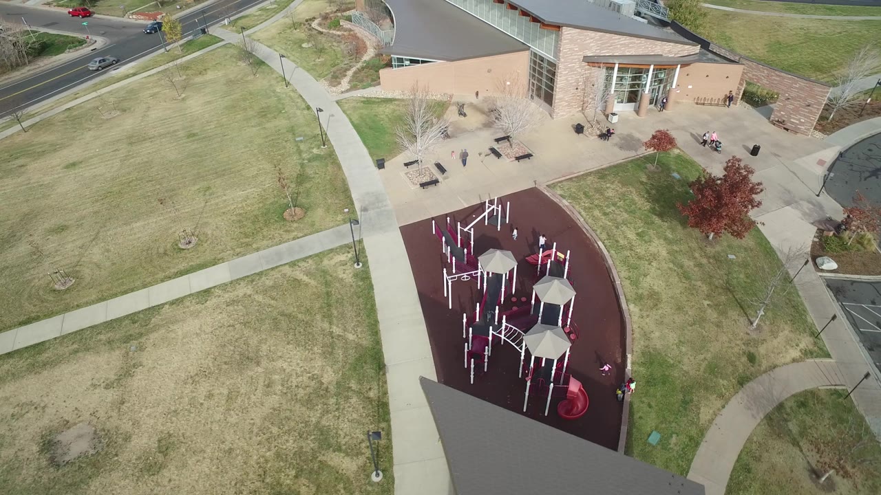 A second overhead view in Hidden Lake, Colorado centers the red playground surface below the yellow building, with open grass, crossing sidewalks, parked benches, street traffic, and small visible people.