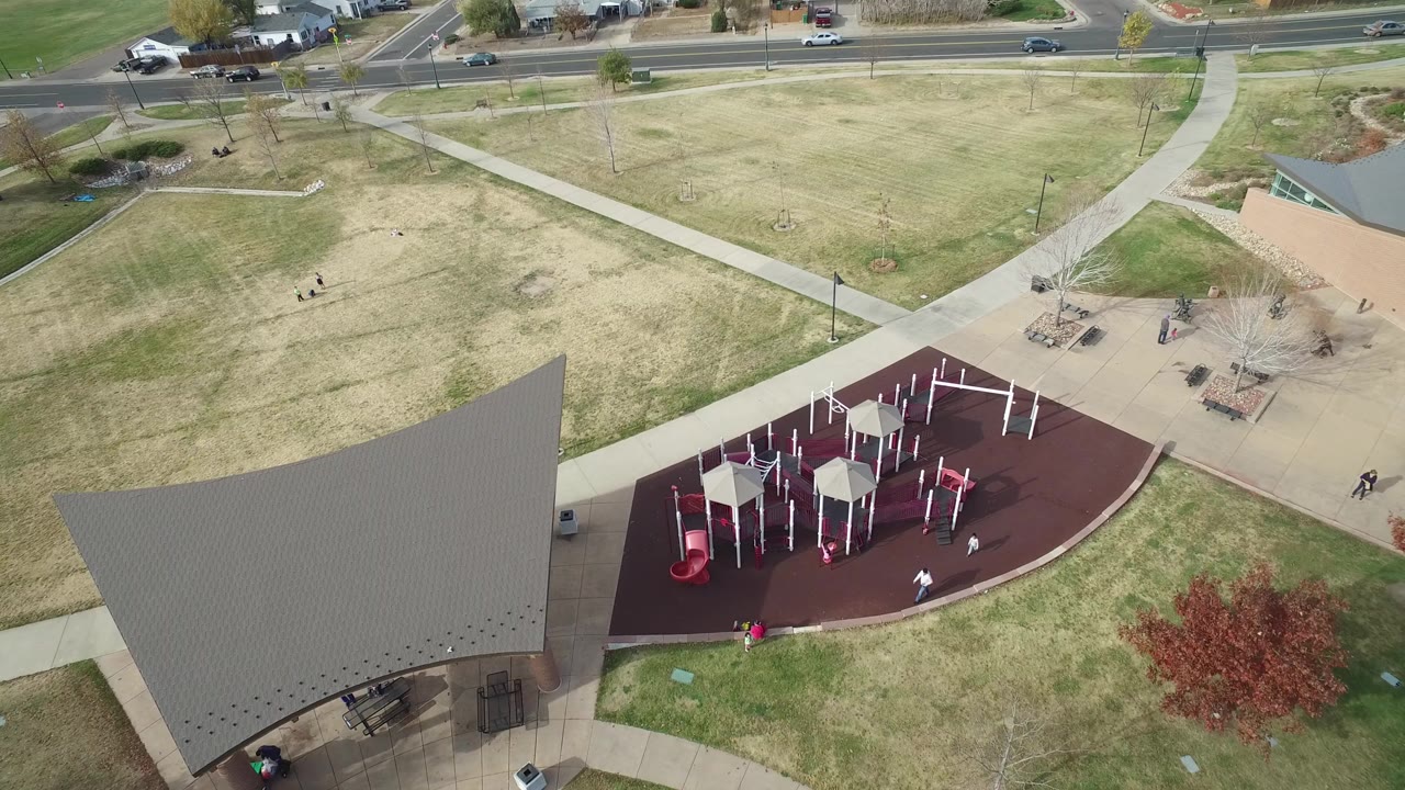 Aerial frame over Hidden Lake, Colorado showing a red playground surface, large angular shade roof, concrete paths, open grass, roadside traffic, and small people visible below.