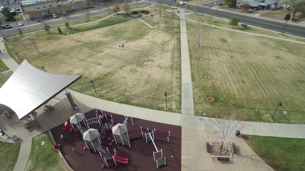 High-angle view of a Hidden Lake, Colorado park with a white canopy tent at lower left, playground equipment at lower center, crossing paths, dry grass, roads, and small people visible in the field.
