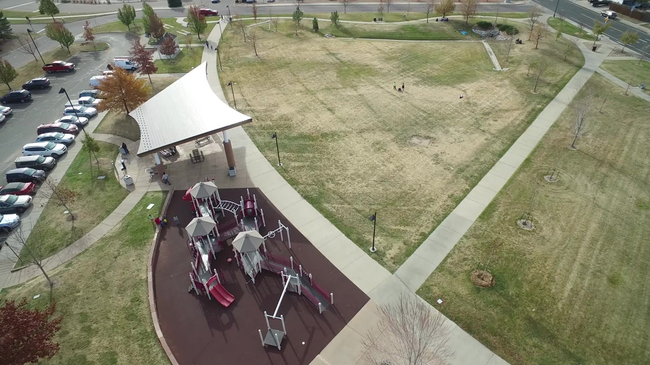 Aerial view of a playground, pavilion, paths, parked cars, and open grass.