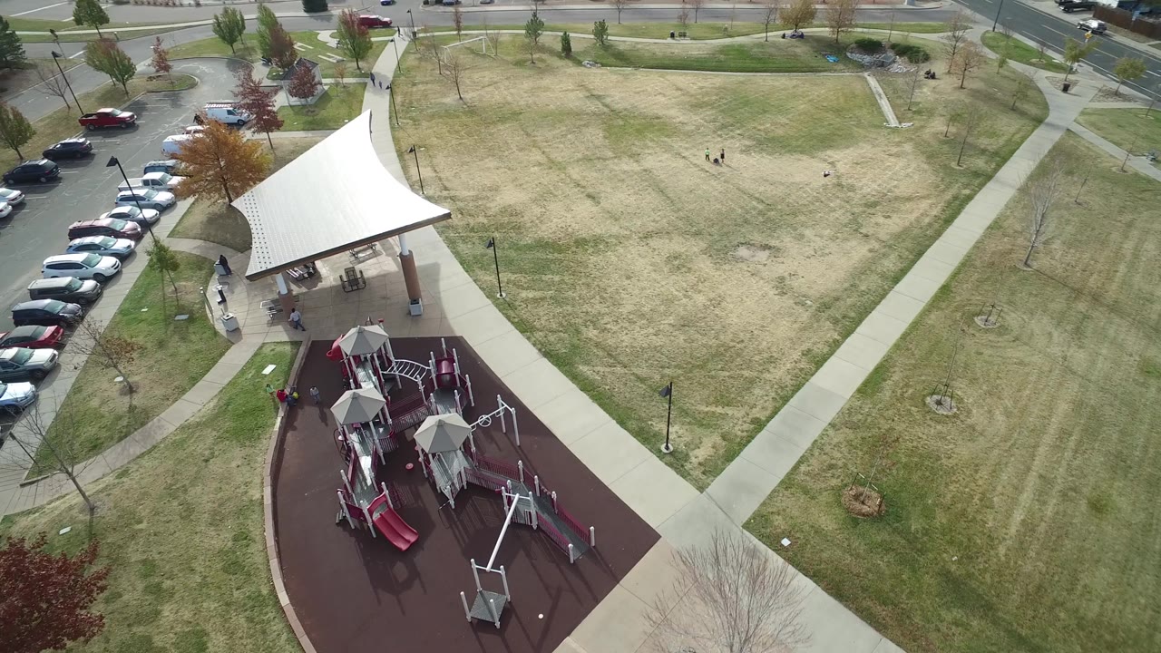 Aerial view in Hidden Lake, Colorado with the playground closer to center, the pavilion beside a curved path, rows of parked cars along the road, and small distant people visible in the grass.