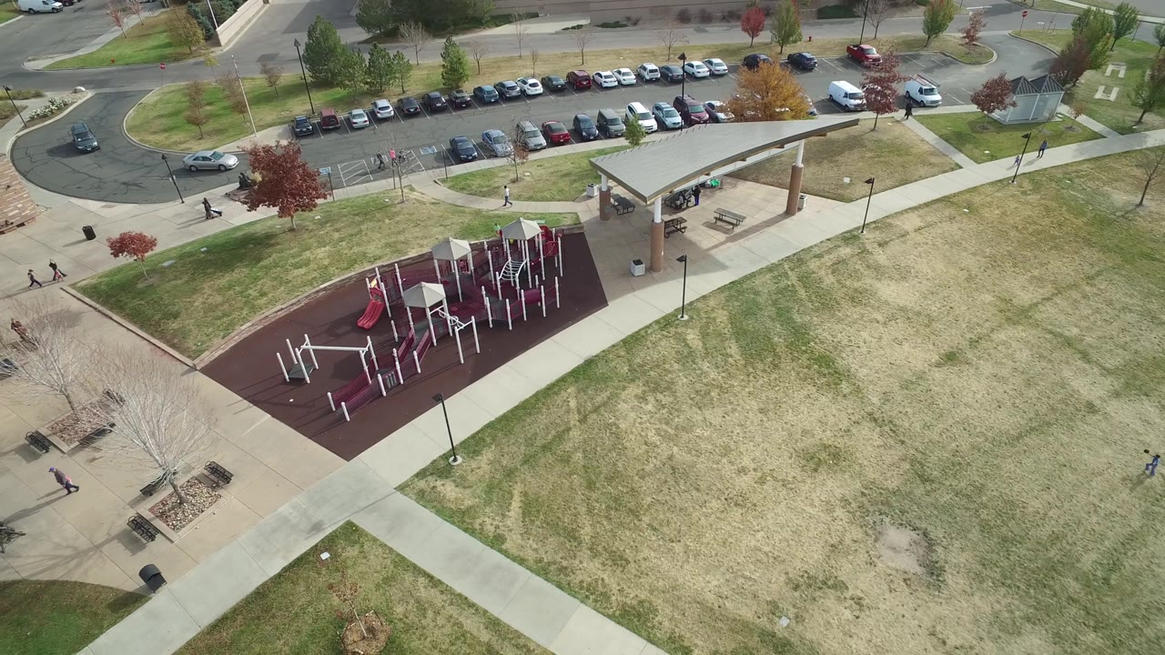 Aerial frame of a red playground pad, shelter, lawns, sidewalks, cars, and curved streets.