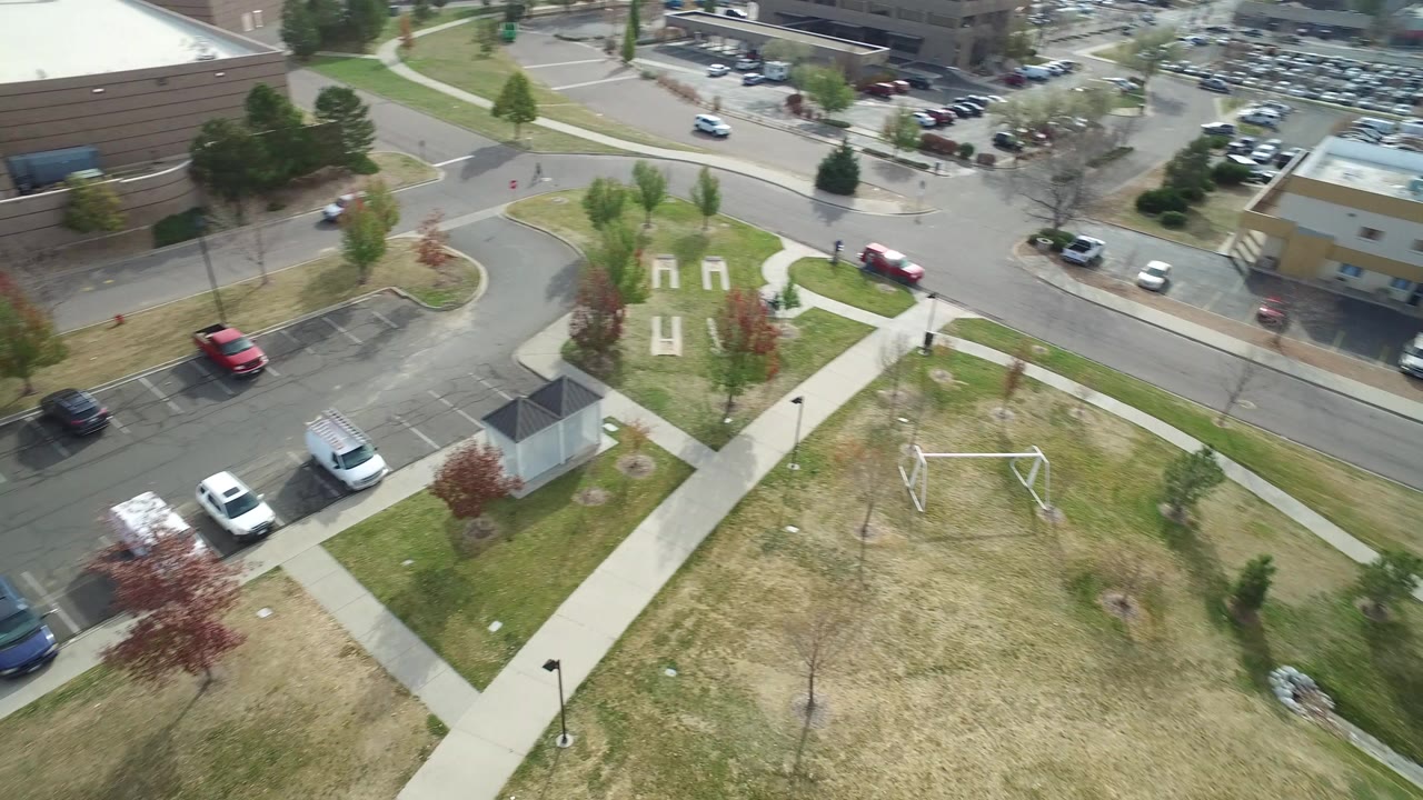 Elevated view of a grassy park with paths, a swing frame, streets, parked cars, and low-rise buildings.