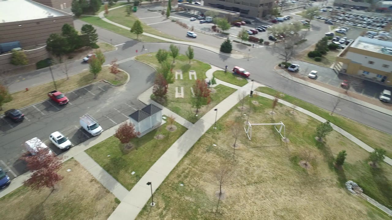 Aerial frame in Hidden Lake, Colorado with the park paths and swing frame below, a red vehicle on the street, parking areas, and a tan low-rise building across the road.
