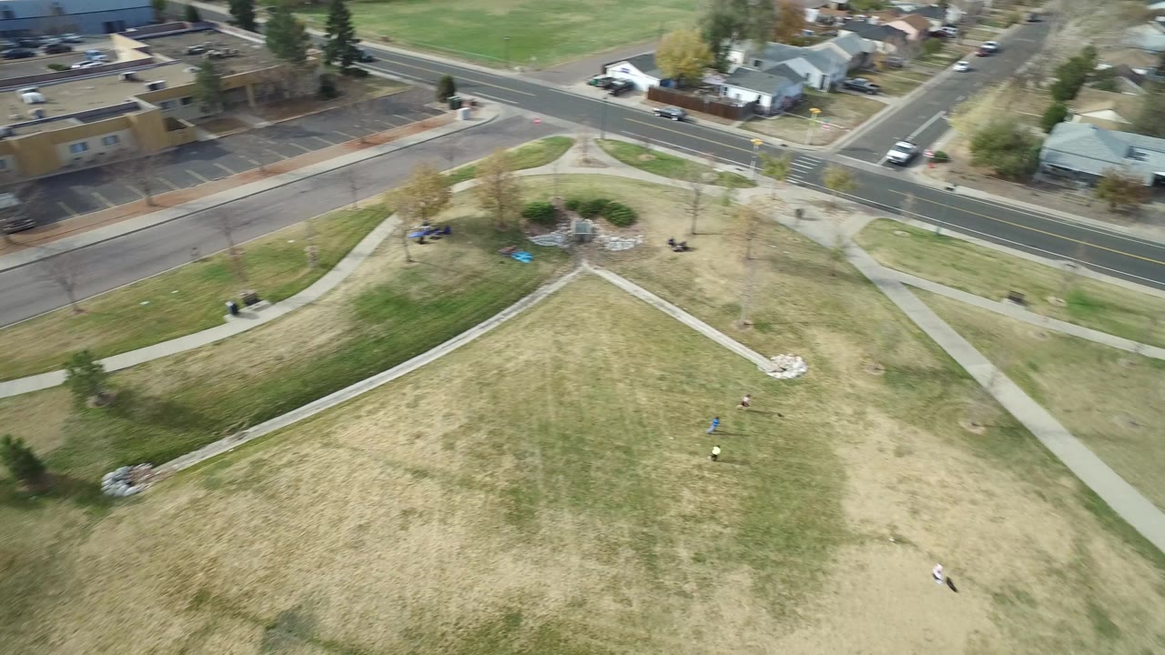 Aerial view of the Hidden Lake, Colorado lot with pale grass, intersecting concrete paths, a road and homes at the top edge, and small trees casting short shadows.