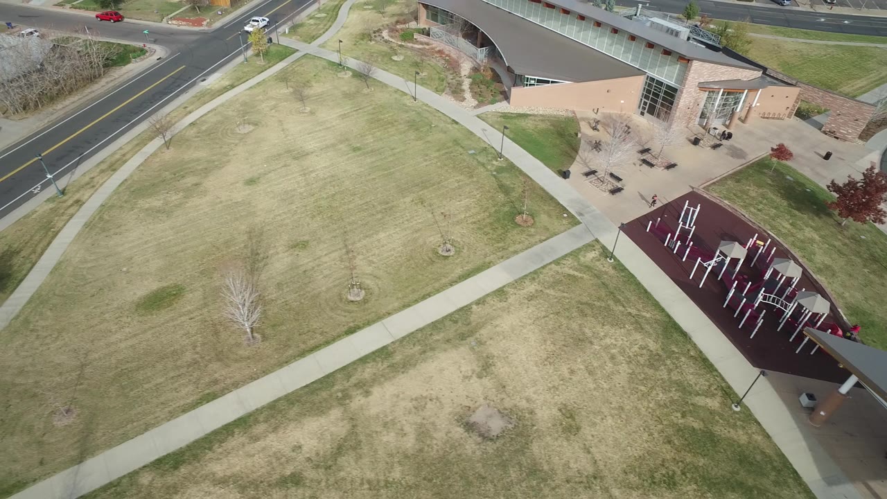 Aerial frame in Hidden Lake, Colorado with the field centered, diagonal walking paths, a two-lane road, a red car, and the building edge at upper right, with no visible people.