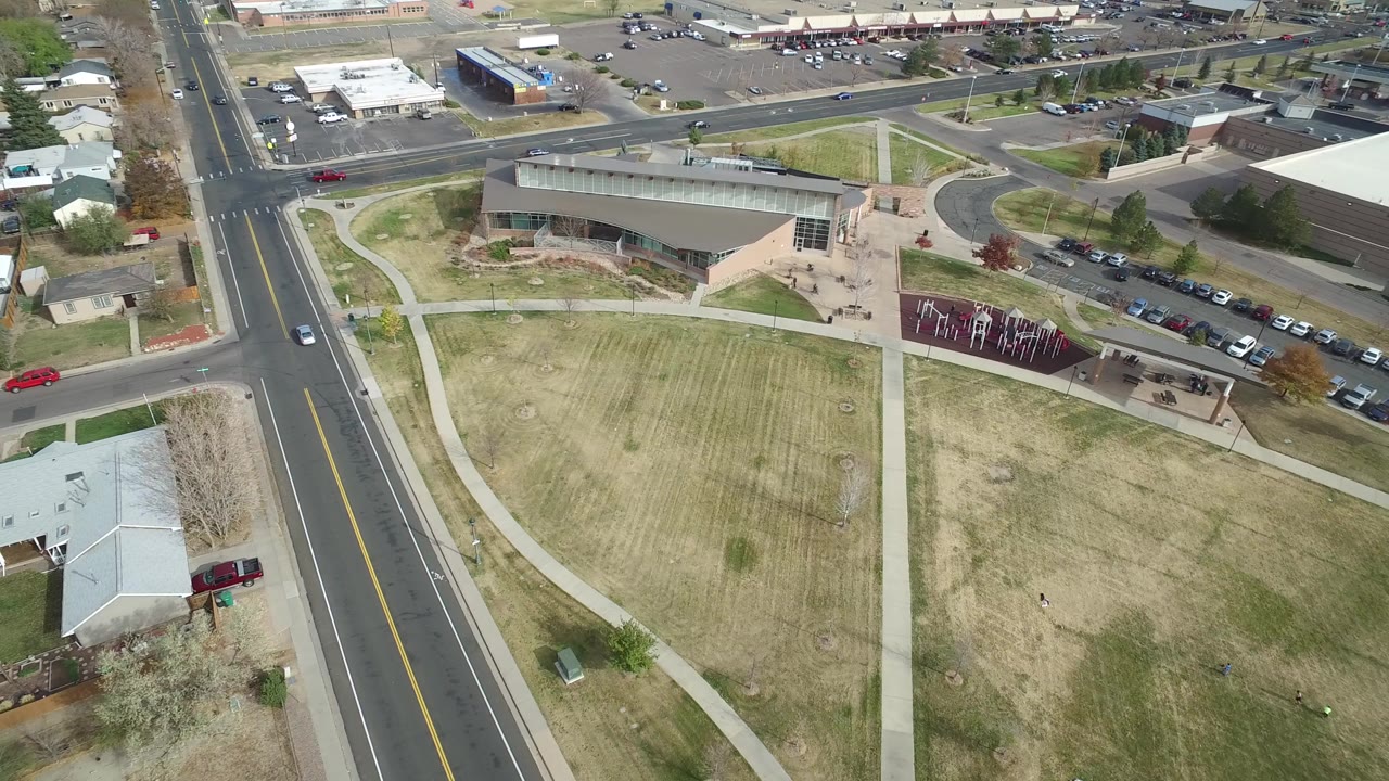 High aerial frame over Hidden Lake, Colorado showing a multi-lane road, pale lawn, crossing sidewalks, parked cars, and low-rise buildings with no visible people.