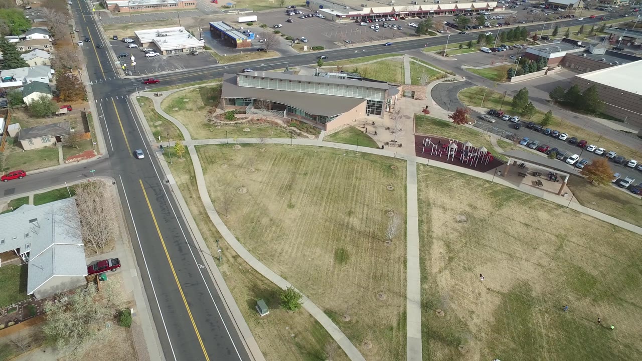 Aerial view in Hidden Lake, Colorado with the road running down the left side, a large mowed lawn in the middle, sidewalks, parking spaces, and commercial or civic buildings beyond, with no visible people.