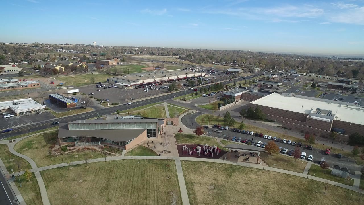 Wide aerial frame over Hidden Lake, Colorado, showing a large low building, parking areas, curved sidewalks, lawns, roads, distant houses, and no visible people.
